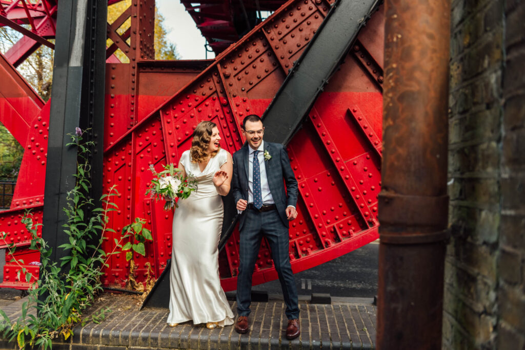 Couples dancing in front of a red and black iron bridge, the bride wears a cream cowl neck on the byas silk dress and has Hollywood waves in her hair and the groom wears a navy suit and navy tie bride holds a small bouquet of cream roses and green foliage