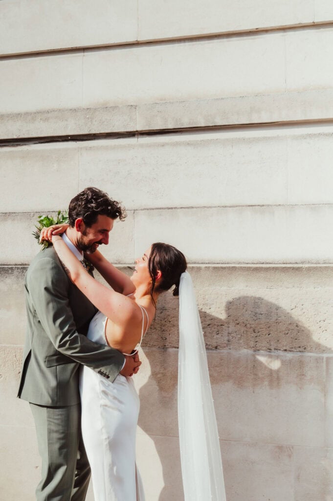 Bride in cream Reformation dress with long veil and gold shoes laughing with groom in sage green suit outside Hackney Town Hall