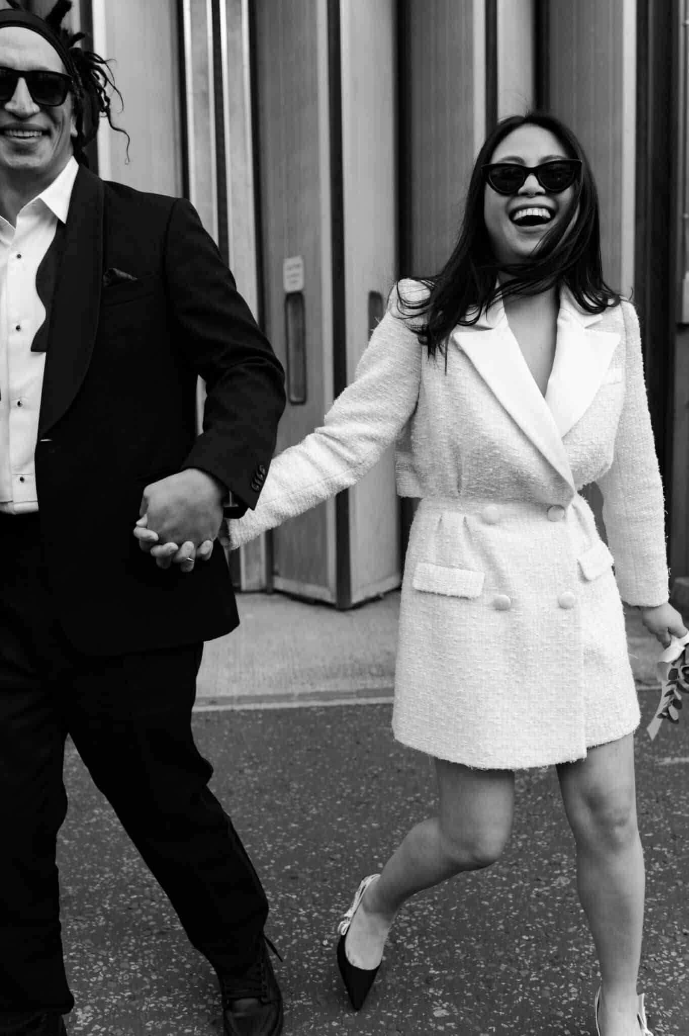 Couple leaving Islington Town Hall, bride in a cream Self-Portrait suit jacket dress and J’adore Dior heels, groom in a Buckleigh London tuxedo with a loose bow tie and Cartier rings.