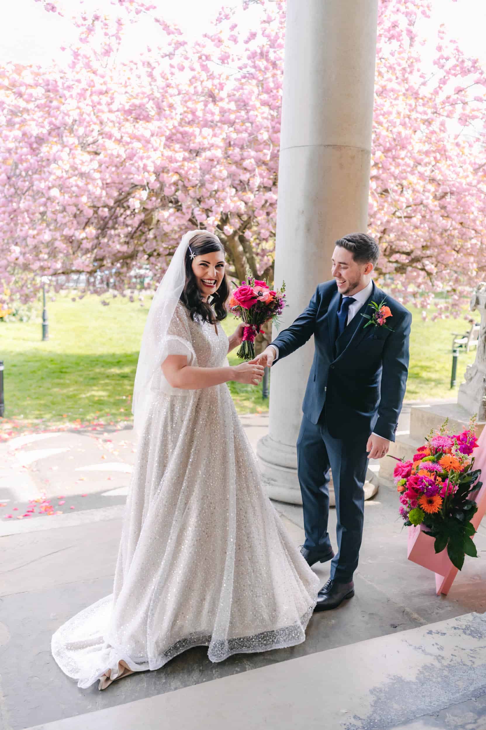 Joyful couple laughing during spring wedding at The Asylum Peckham, bride in sparkle dress with cherry blossom backdrop, candid London wedding photographer
