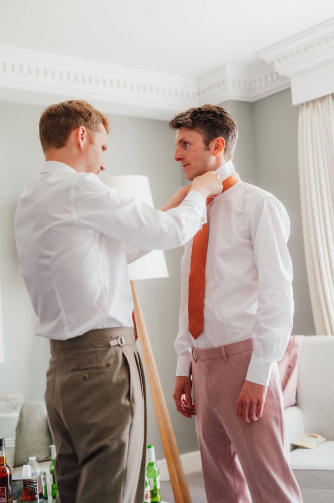 Groom and best man getting ready for wedding, adjusting ties, stylish clothing, elegant interior setting.
