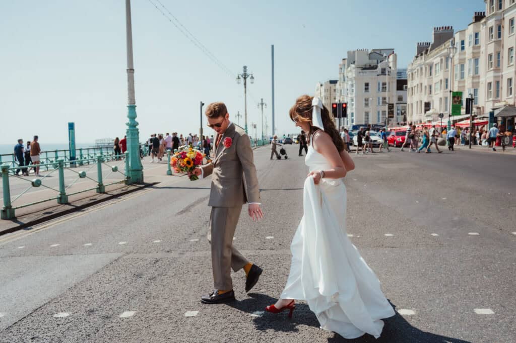 Walk along seaside wedding photo of bride and groom on a sunny day in Brighton, with vibrant city background and onlookers.