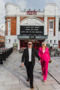 Couple in colourful formal wedding outfits walking hand in hand outside The Ritzy Cinema in Brixton
