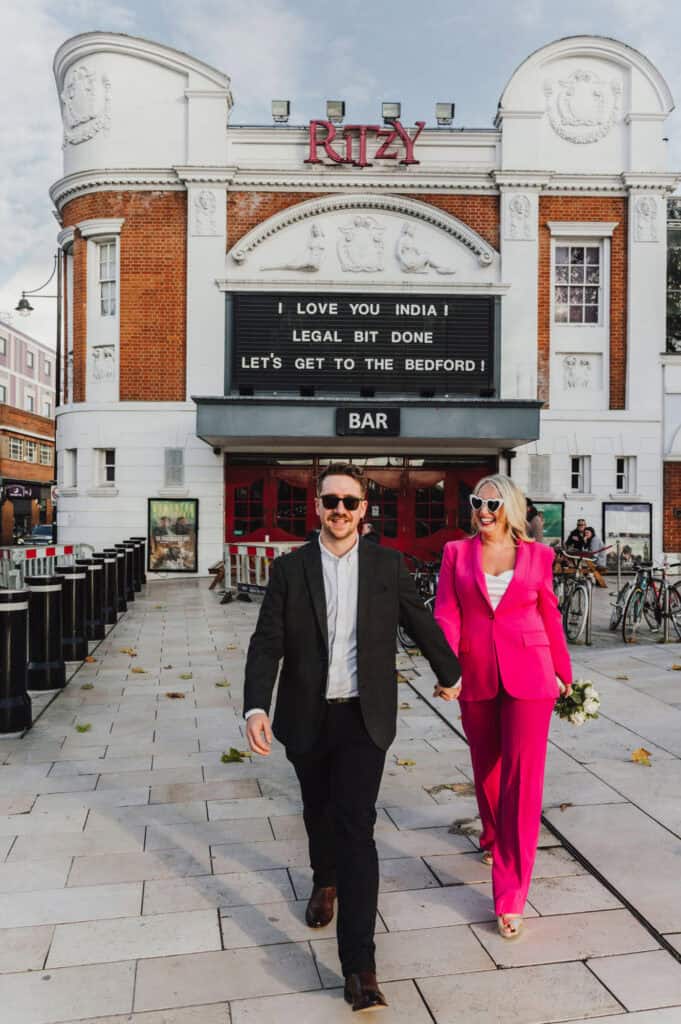 Couple in colourful formal wedding outfits walking hand in hand outside The Ritzy Cinema in Brixton