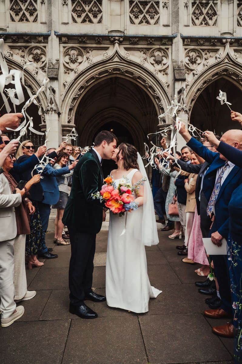 Couple kissing outside church with guests waving streamers and colourful bouquet, London wedding by Sidey Clark