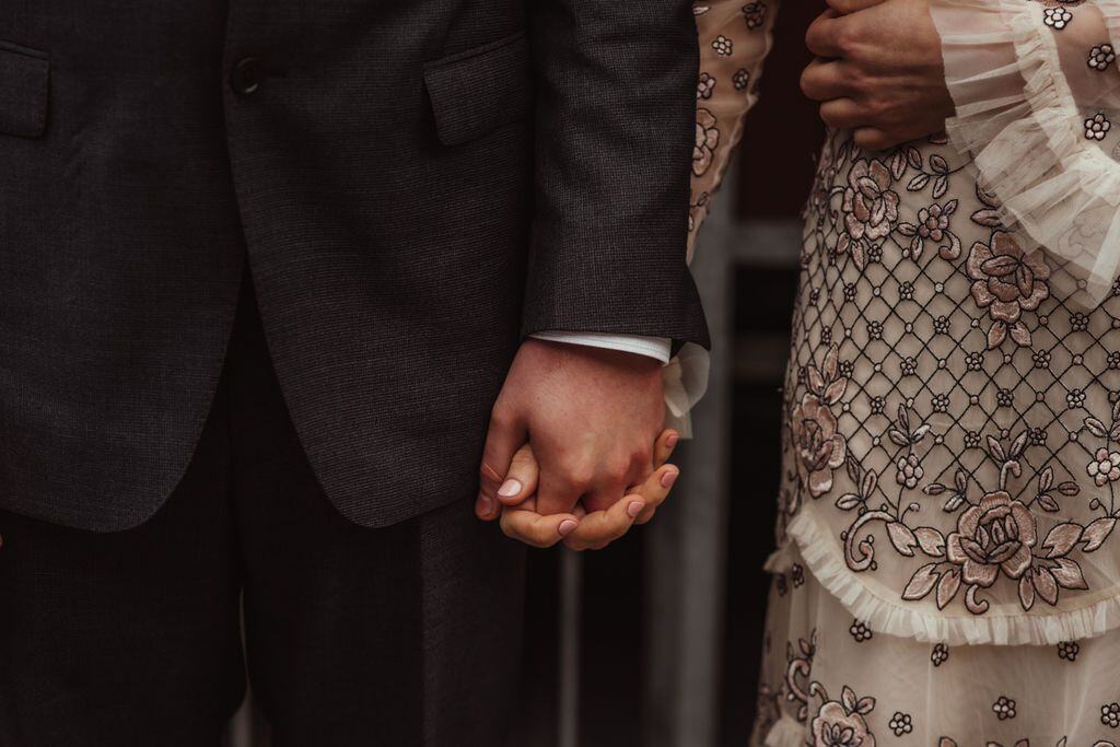 Close-up of couple holding hands, bride in embroidered lace dress, intimate wedding detail by Sidey Clark