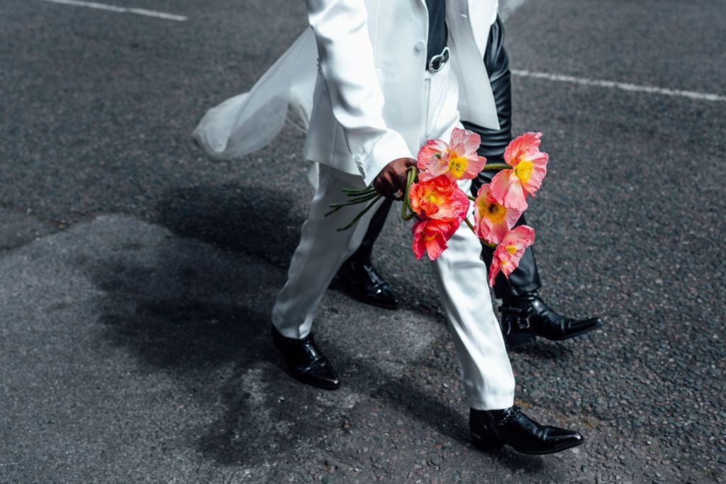 Groom in white suit walking with pink poppies on a London street, documentary wedding photography by Sidey Clark