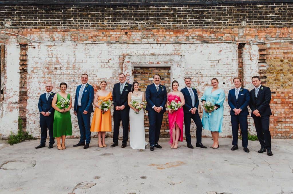 Wedding party lined up against an exposed brick wall in colourful outfits, London wedding by Sidey Clark